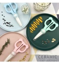 A blue Cubble ceramic food scissors rests on a blue food plate, a pink ceramic food scissors rests on a pink plate, a green ceramic scissors rests on a green plate. They are surrounded by cut food