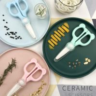 A blue Cubble ceramic food scissors rests on a blue food plate, a pink ceramic food scissors rests on a pink plate, a green ceramic scissors rests on a green plate. They are surrounded by cut food